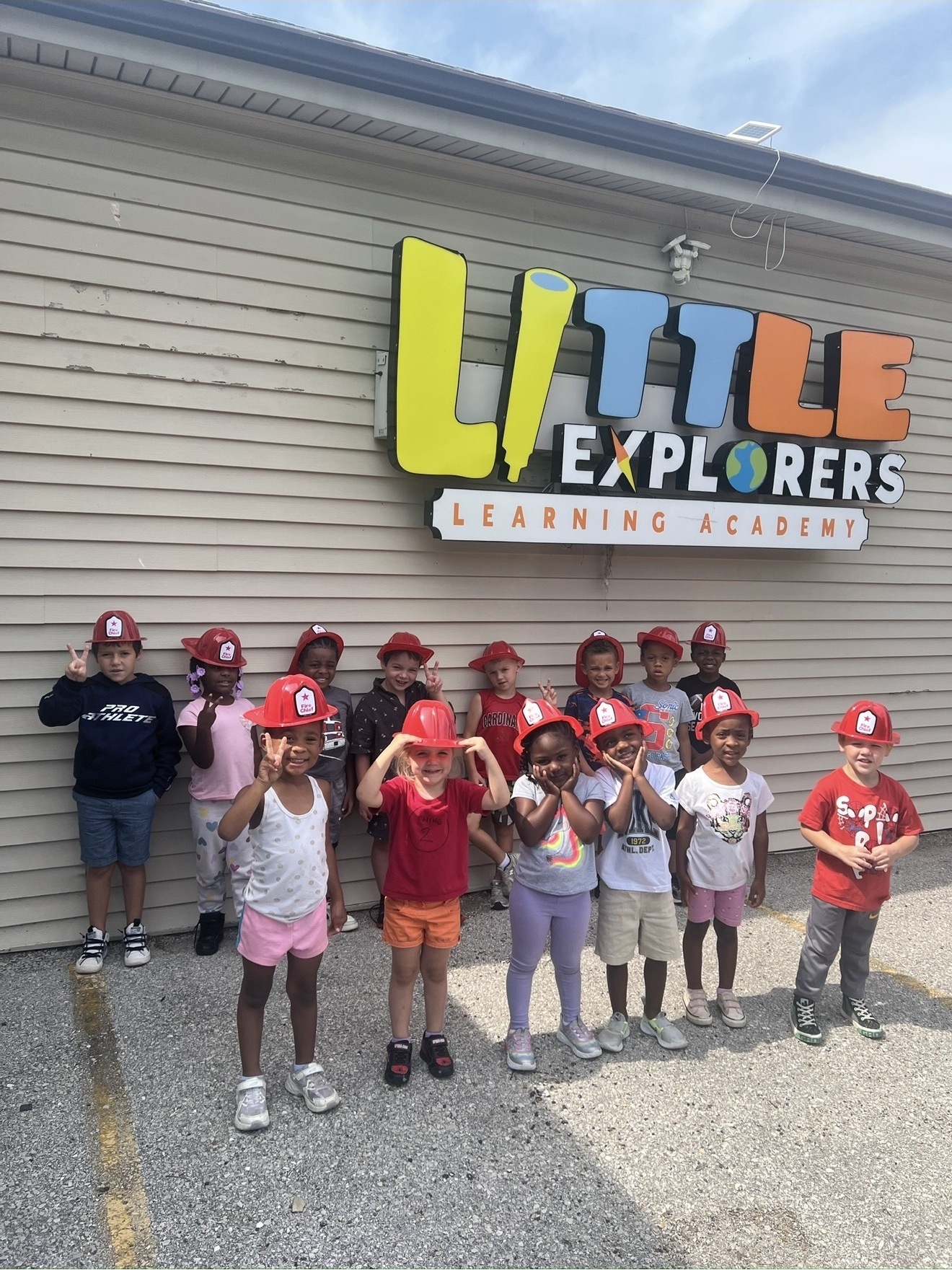 Children from Little Explorers Learning Academy lined up outside in red firefighter helmets, in front of the school sign