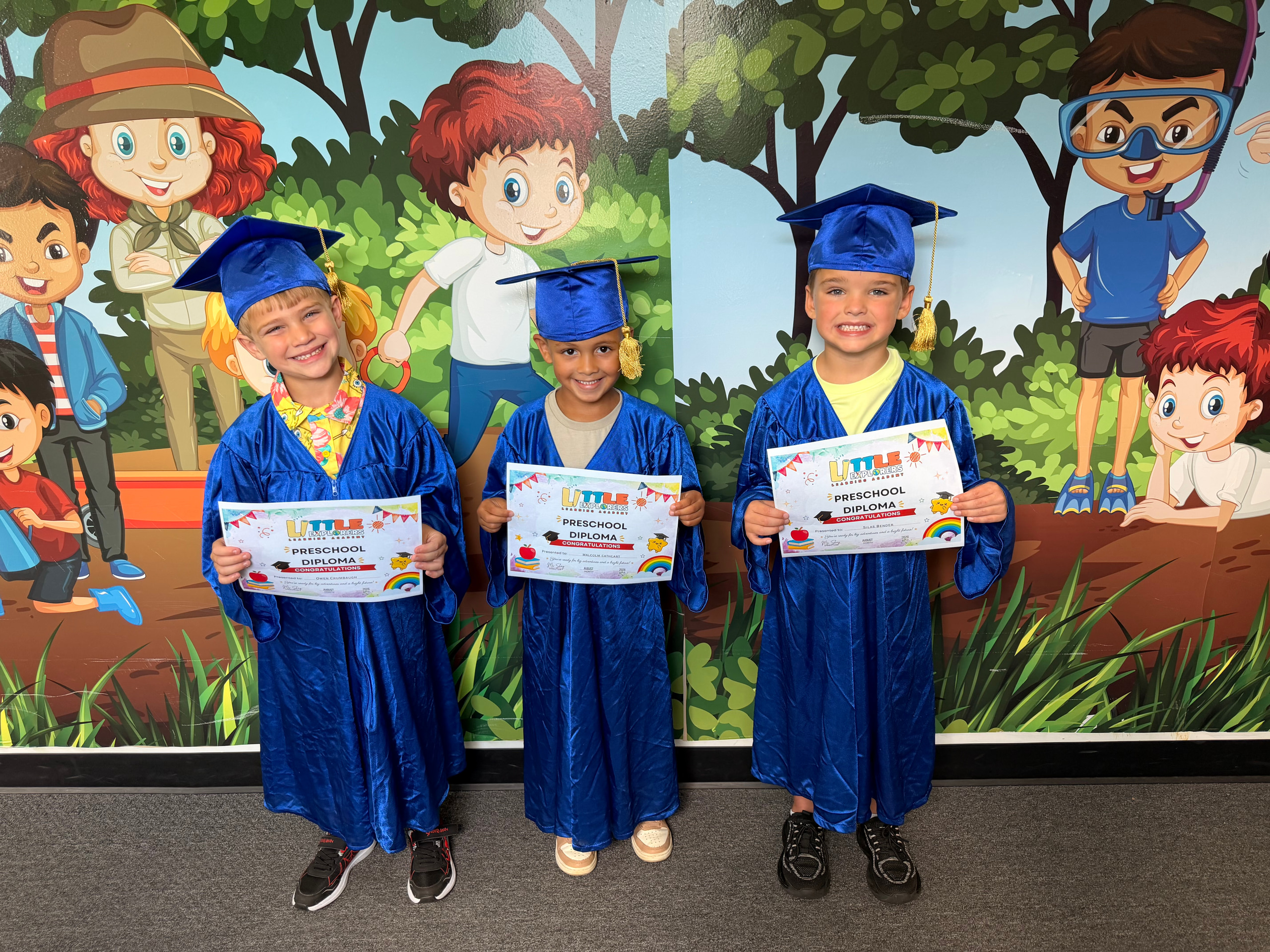 Three Little Explorers graduates in blue caps and gowns holding their preschool diplomas in front of the classroom mural